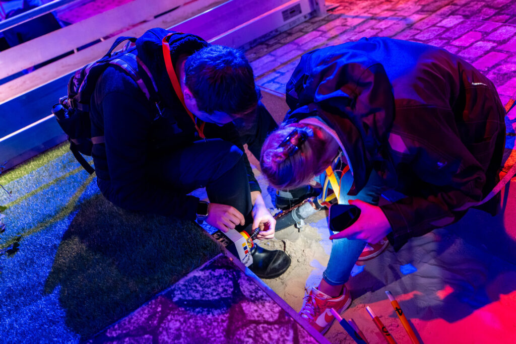 Drie deelnemers zoeken aanwijzingen in zand onder gekleurd licht.