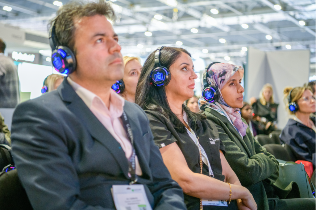 3 people are seated watching a live conference session at CCR London. They are all wearing badges and lanyards and wearing LED headphones