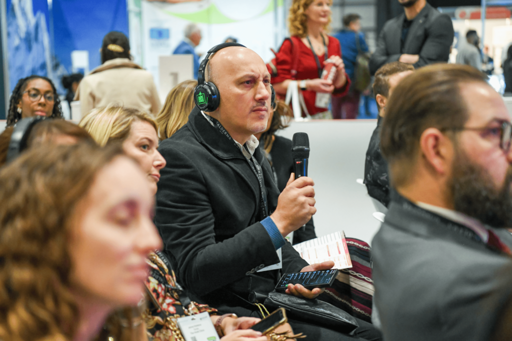 A mean is seated within a conference audience, they are all wearing headphones and he is holding a microphone asking a question