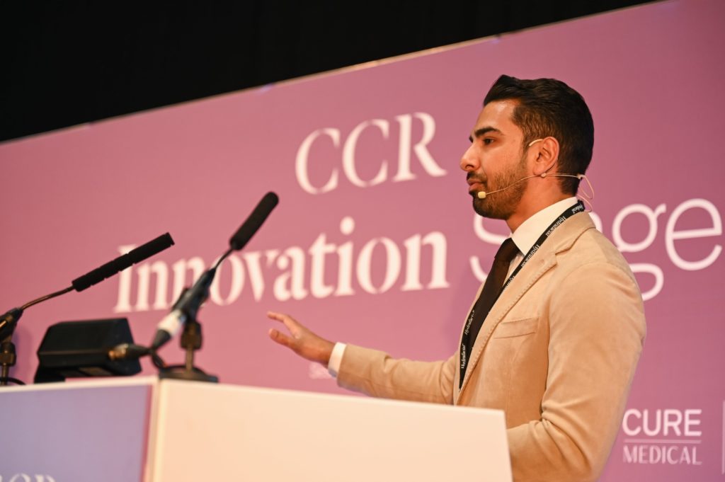 Speaker talking on the innovations stage at CCR 2024. he is wearing a light pink suit standing in front of a purple backdrop