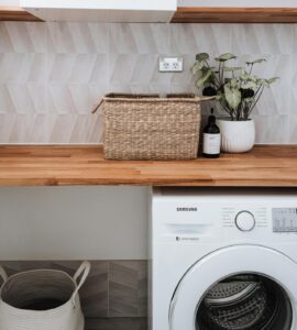Close-up van een wasruimte met een wasmachine, een werkblad en een paar wandplanken. Naast de wasmachine staat een wasmand. Op het werkblad staan een mand, zeep en een plant.