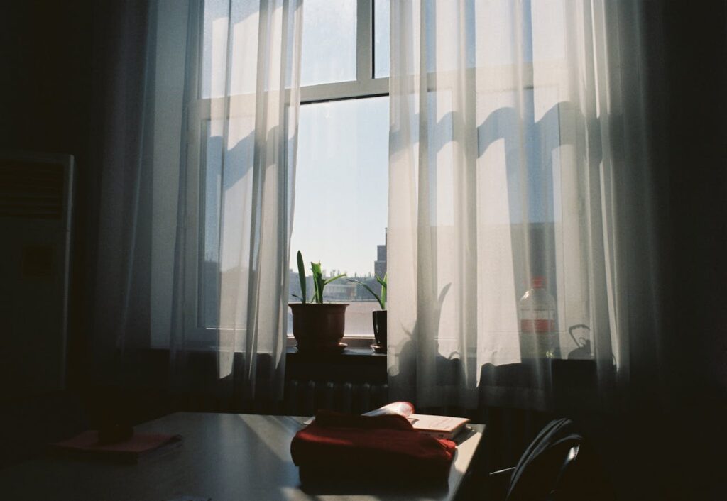 Donkere afbeelding van een raam met blauwe lucht en daarvoor witte, semi-transparante vitrages.