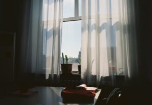 Donkere afbeelding van een raam met blauwe lucht en daarvoor witte, semi-transparante vitrages.