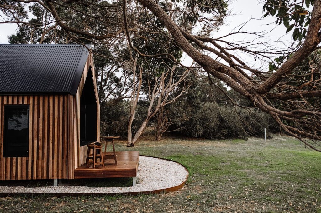 Een tiny huis van hout met een terras. Er bevinden zich twee stoelen en een tafel op het terras. Op de achtergrond zie je een groot grasperk en bomen en struiken.
