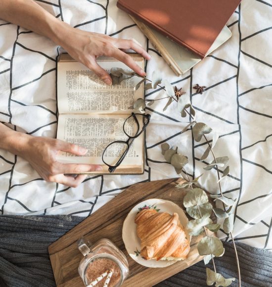 Een bed met daarop een paar boeken, iemand die ene boek openhoudt, een leesbril op het geopende boek en een houten plank met een croissant en een chocolademelk erop.