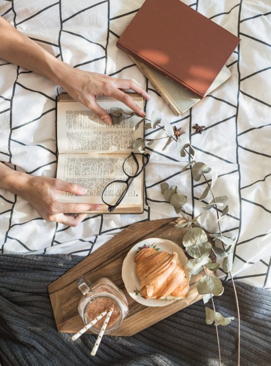 Een bed met daarop een paar boeken, iemand die ene boek openhoudt, een leesbril op het geopende boek en een houten plank met een croissant en een chocolademelk erop.