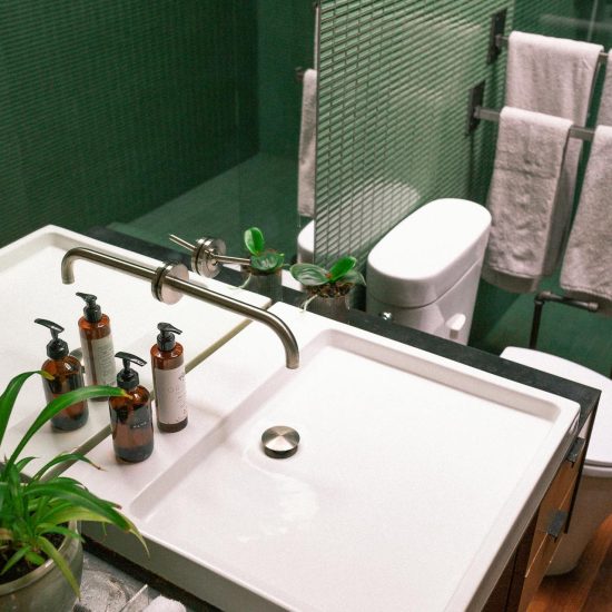 Close-up afbeelding van een badkamer met groene tegels met de focus op een lavabo met twee zeeppompjes en een plantje.