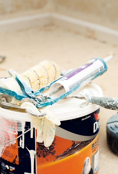 Hoek van een badkamer met werkmateriaal op de grond. Waaronder een pot verf, voegkit, verfrollers, borstels, enzovoort.