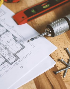 Een afbeelding van de plannen van een gebouw met daarnaast een meetlint, boormachine, waterpas en een paar vijzen.