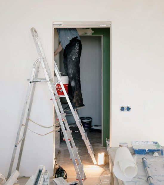Een ruimte met een man die achter een deuropening schilderwerken uitvoert op een ladder. In de ruimte staat ook allerlei renovatiemateriaal zoals een radiator, werktafel en schildergerief.