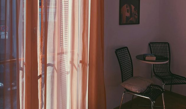 Afbeelding van een hoek van een kamer met twee stoelen en daarnaast een raam met oranjeroze vitrages.