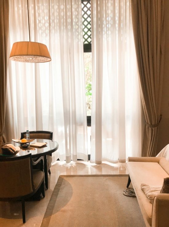 Lichte woonkamer in beigetinten met centraal een raam met witte vitrages ervoor. Naast het raam bevinden zich een zetel en een tafel met twee stoelen.