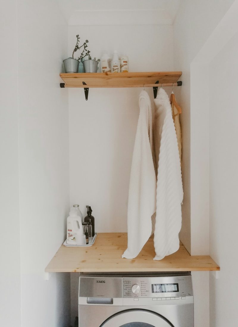 Een kleine wasruimte met een wasmachine met daarop een houten werkblad. Daarboven hangt een plank met daaronder een stang met een paar kapstokken en kledingsstukken. Op het werkblad bevindt zich zeep.
