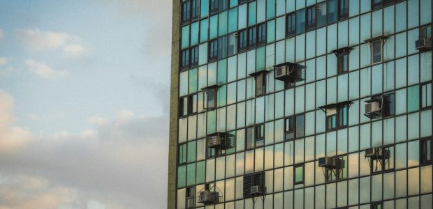 Een ingezoomde hoek van een grote wolkenkrabber die enkel uit glas bestaat. Een paar ramen staan open, aan een paar ramen hangen airco/ventilatiesystemen.