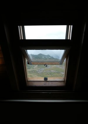 Een donkere afbeelding met een schuin dakraam met uitzicht op de bergen.