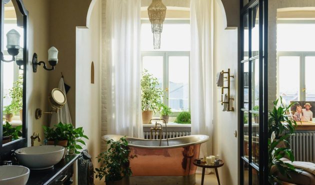 Vintage, luxueuze badkamer met een koperen badkuip die voor een groot raam met vitrages staat. Een de linkerkant bevinden zich lavabo's met een grote spiegel. De muren zijn olijfgroen geverfd.