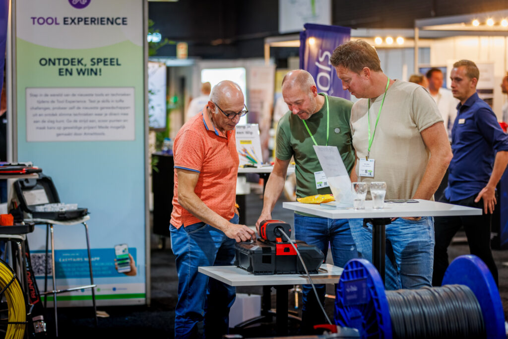 Drie bezoekers bekijken een demonstratie van gereedschap aan een tafel.