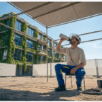 Installatiemonteur drinkt water op een bouwplaats onder een overkapping tijdens een hete zomermiddag, met felblauwe lucht, moderne gebouwen met groene gevels op de achtergrond.
