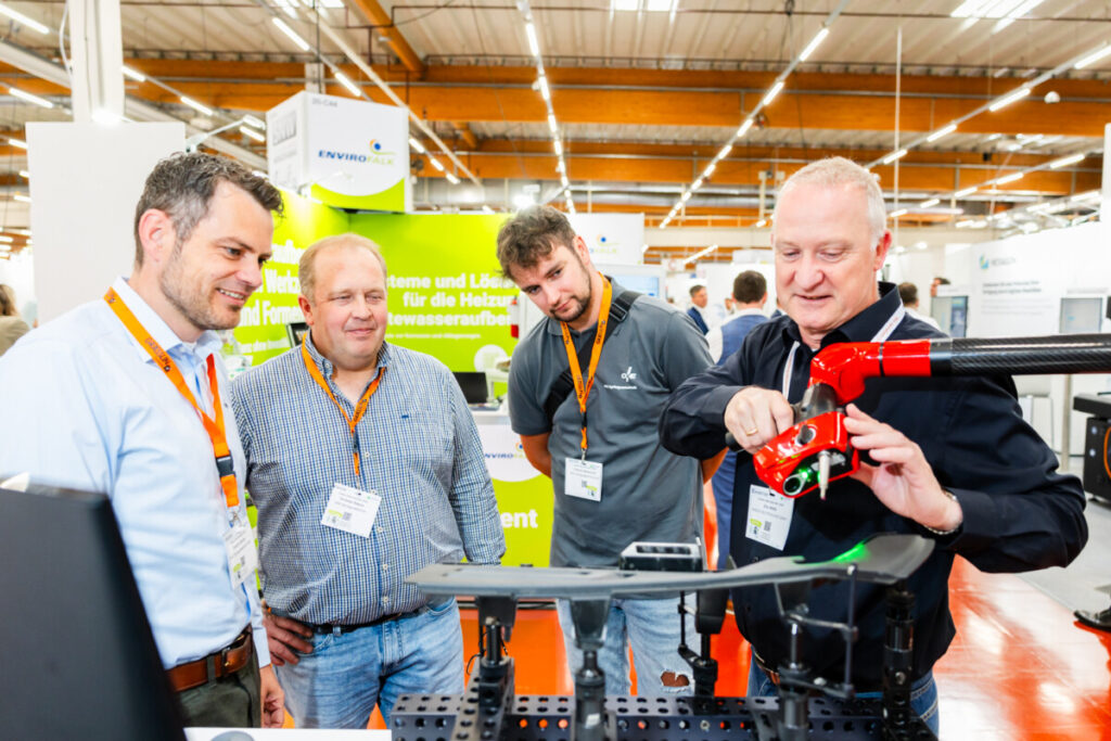 KUTENO Messe 2025: 4 Männer reden über einen Roboterarm in ihrer Mitte