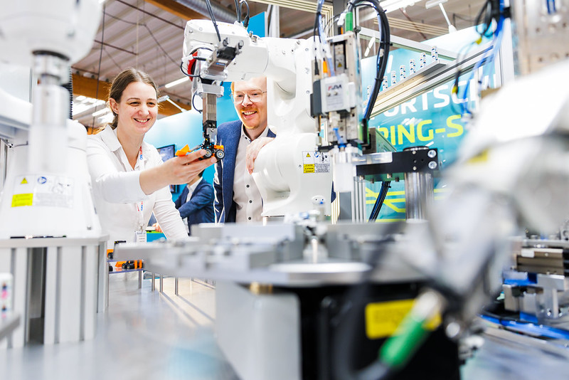 Zwei Personen stehen auf der KUTENO / KPA in Bad Salzuflen an einer automatisierten Fertigungsanlage mit Industrierobotern; im Vordergrund sind unscharf Greifarme und F&ouml;rdertechnik zu sehen, w&auml;hrend eine Person im Hintergrund ein Bauteil in der Hand h&auml;lt und die Technik erkl&auml;rt.