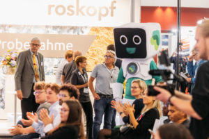 Publikum ist von vorne fotografiert und klatscht gerade, das Messe-Maskotchen der Badgy klatscht auch