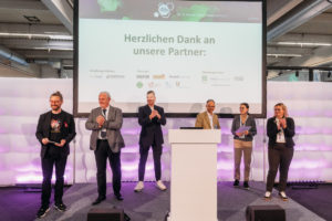 Die Jury steht auf der Bühne und verkündet die Gewinner, wir sehen sie frontal vor der LEinwand stehend