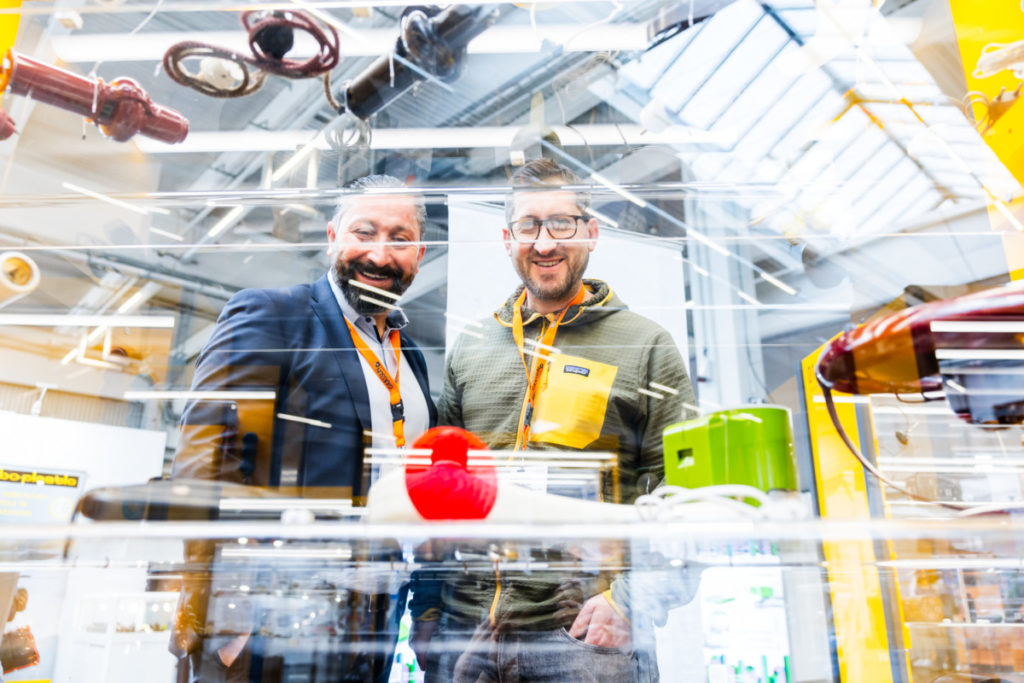 Zwei lachende Männer, die durch eine Vitrine hindurch fotografiert wurden, auf deren Glasboden Kunststoffprodukte liegen.