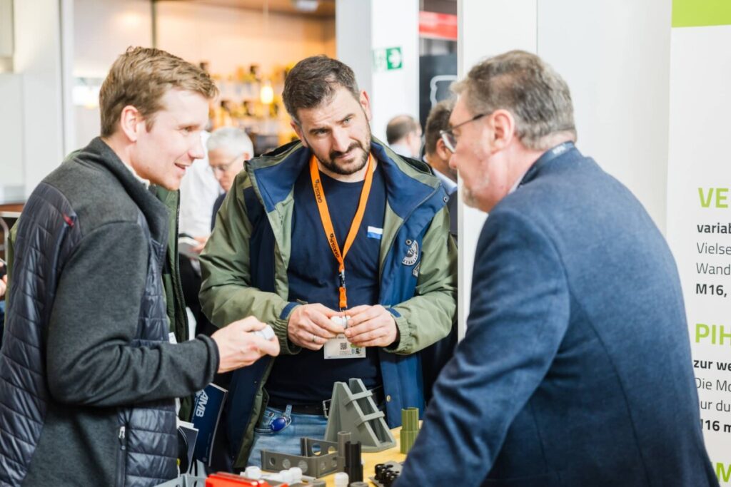 Drei M&auml;nner stehen auf der KPA in Ulm 2026 an einem Messestand und sprechen miteinander. Auf dem Tisch liegen verschiedene Kunststoffteile und Muster.