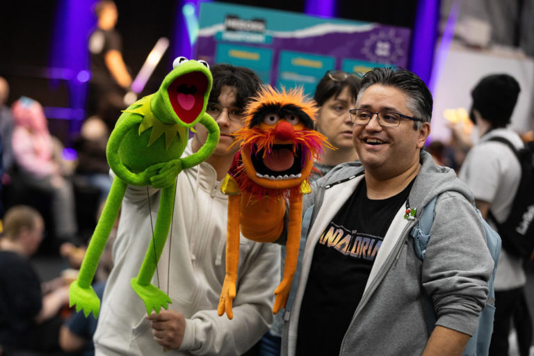 Two hand puppets from the Muppets show, Kermit and Animal, held by two happy Comic Con guests in a crowd.