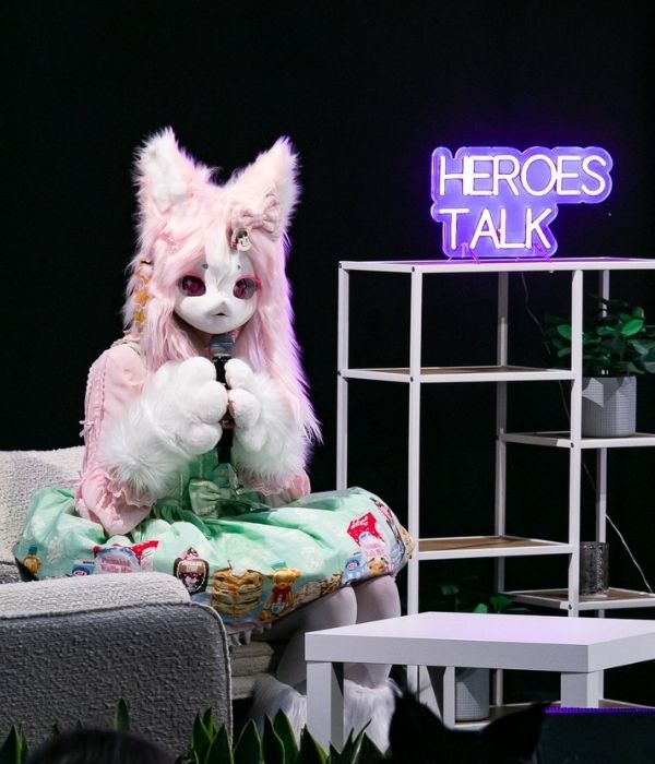 Sabi Mango sitting at the heroes talk stage hosthing a panel. She is dressed in a pink fox mask in colorful pastel clothing.