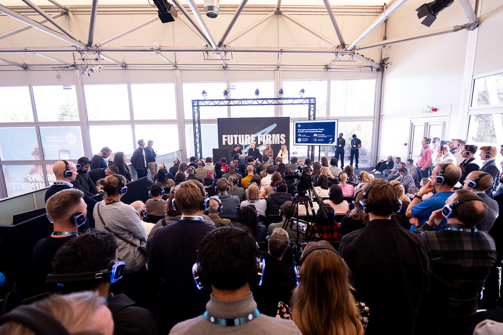 A large audience at an event, with two speakers presenting in the distance