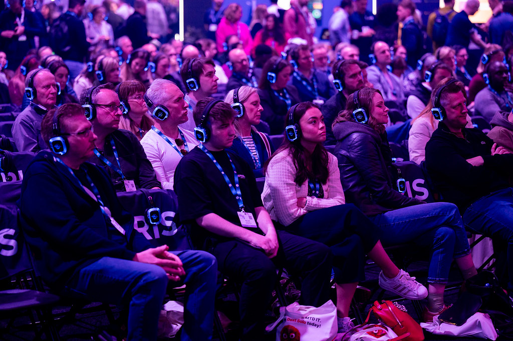 A large audience, all wearing headphones, watching a speaker