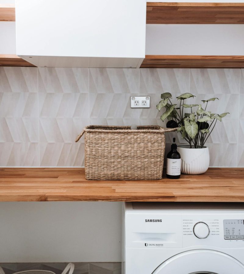 Close-up van een wasruimte met een wasmachine, een werkblad en een paar wandplanken. Naast de wasmachine staat een wasmand. Op het werkblad staan een mand, zeep en een plant.