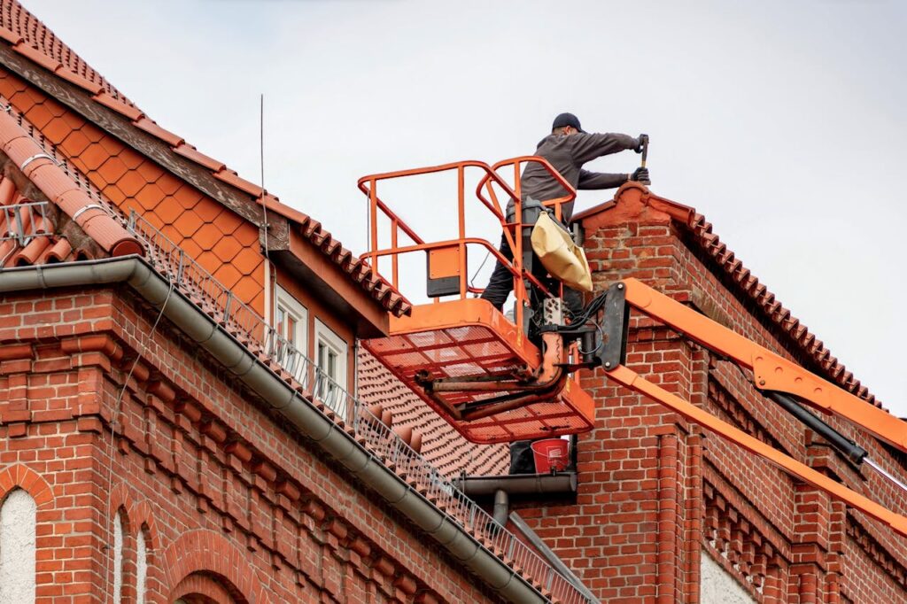 Een dakwerker op een lift die reperatiewerken aan het uitvoeren is aan een oranjerood dak van een huis met rode bakstenen.