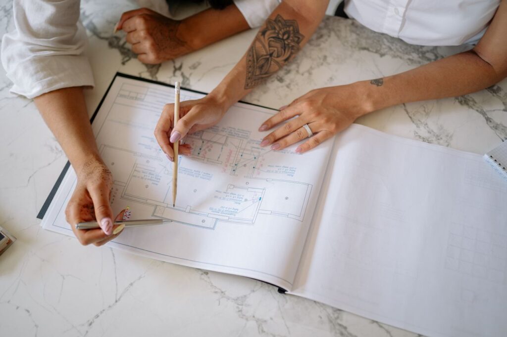 Twee paar handen die met een potlood in de handen uitgebreid naar architecturale plannen kijken.