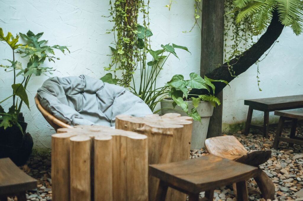 Een terras met grote kiezelstenen met een ronde loungestoel erop. Naast de loungestoel bevinden zich 3 houte nkrukken en 2 cilindervormige bijzettafels.