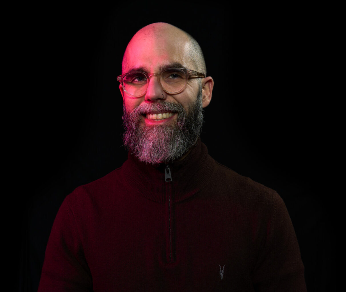 Portrait of Ico Hernandez, wearing glasses and a dark zip-neck jumper against a black background with red lighting.